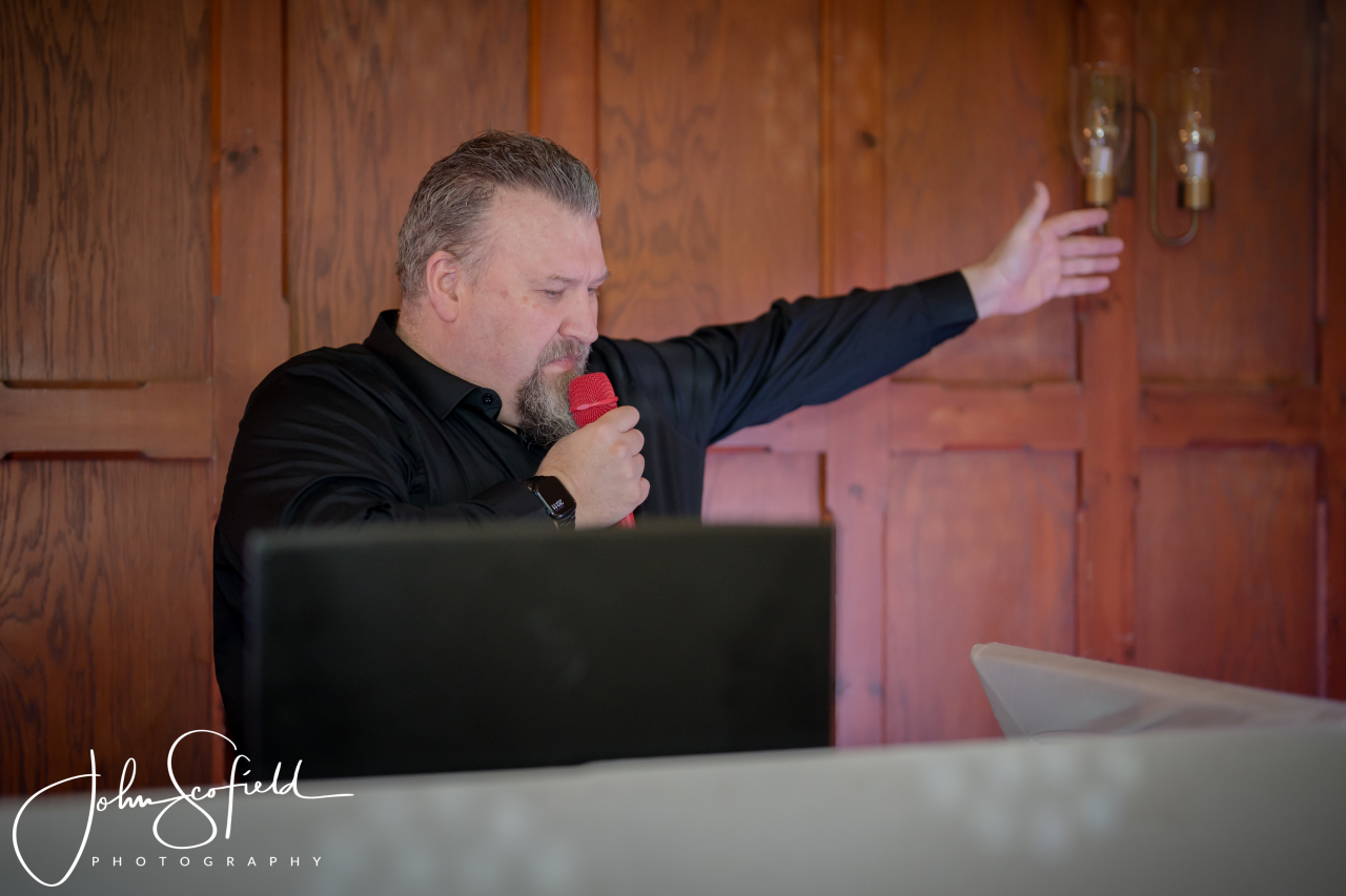 Ian speaking into a red microphone with his other arm extended towards the room